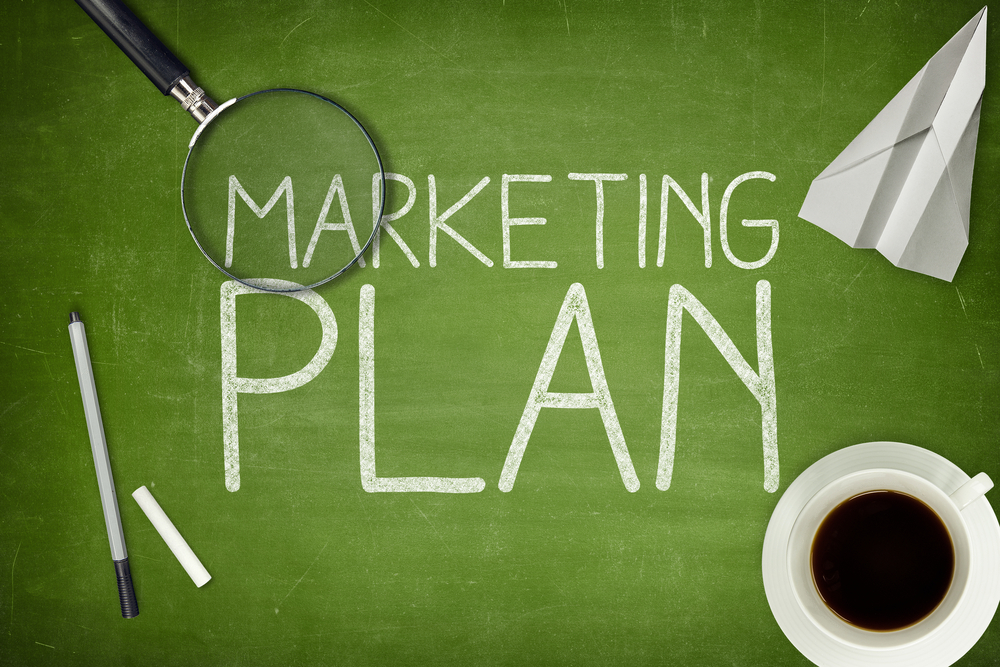 Marketing plan written out on green blackboard. Coffee cup, magnifying glass, chalk, paper airplane and pen resting on blackboard.