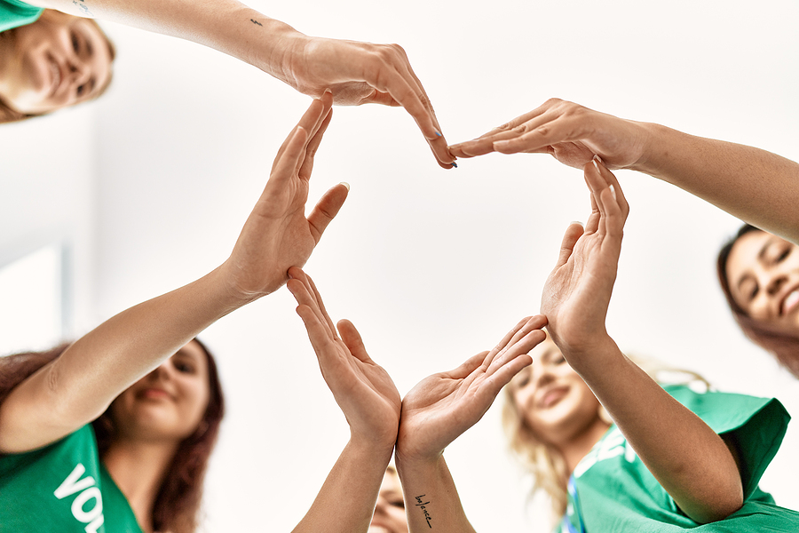 Group of young volunteer women smiling, using joined hands to make heart symbol at charity center